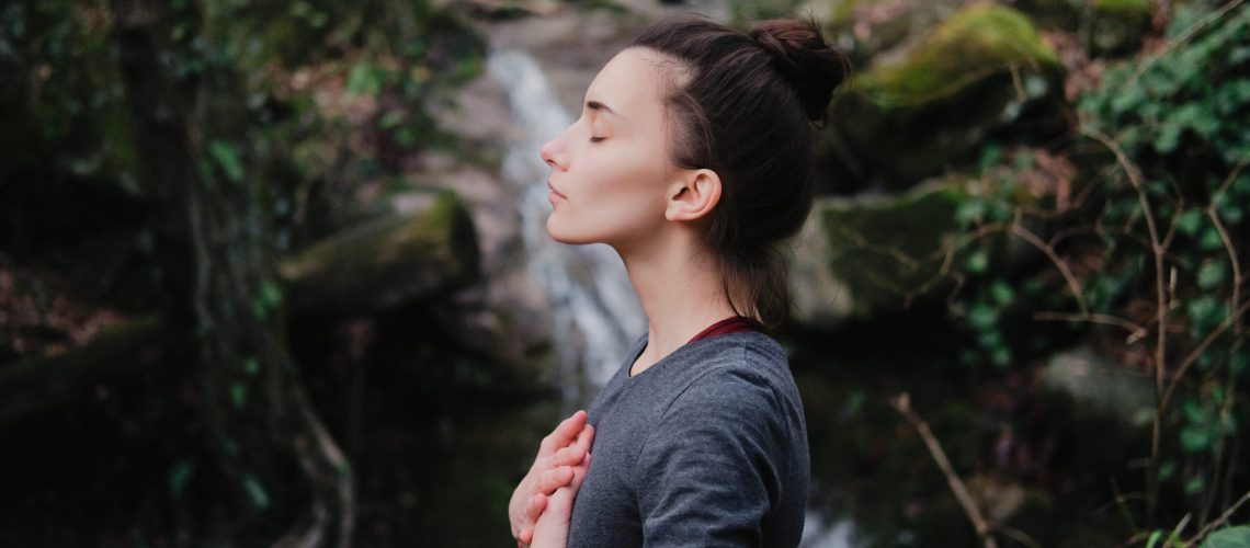 Young woman practicing breathing yoga pranayama outdoors in moss forest on background of waterfall. Unity with nature concept